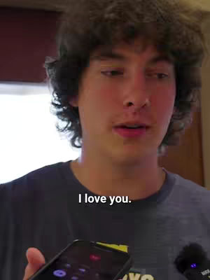 A young man with curly brown hair is shown from the chest up, speaking directly to the camera. He holds a phone in his left hand in front of him. The background is blurred, suggesting an indoors setting. A text overlay reads "I love you."