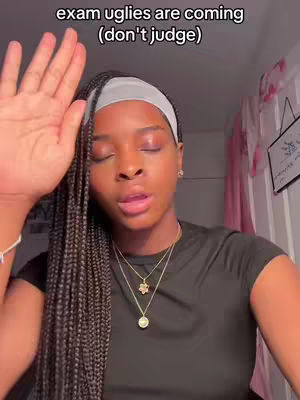 A young Black woman is centered, her upper body filling most of the frame against a softly lit, neutral background. She has long, dark, braided hair and wears a white headband that contrasts with her dark skin. Her eyes are closed, and her lips are slightly parted as if she's mid-sentence or expressing emotion. She's wearing a dark T-shirt and several necklaces, showcasing a delicate, layered jewelry style, the background has soft pink curtains.