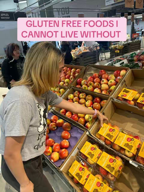 A woman with blonde hair stands in a grocery store in front of a display of apples, picking one. A text overlay reads "GLUTEN FREE FOODS I CANNOT LIVE WITHOUT" in a pink rectangular box.