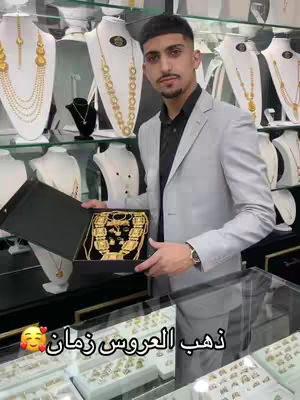 A man in a light gray business suit holds a black jewelry box, displaying an elaborate gold necklace. He stands in a jewelry store, surrounded by showcases filled with gold necklaces on display mannequins.