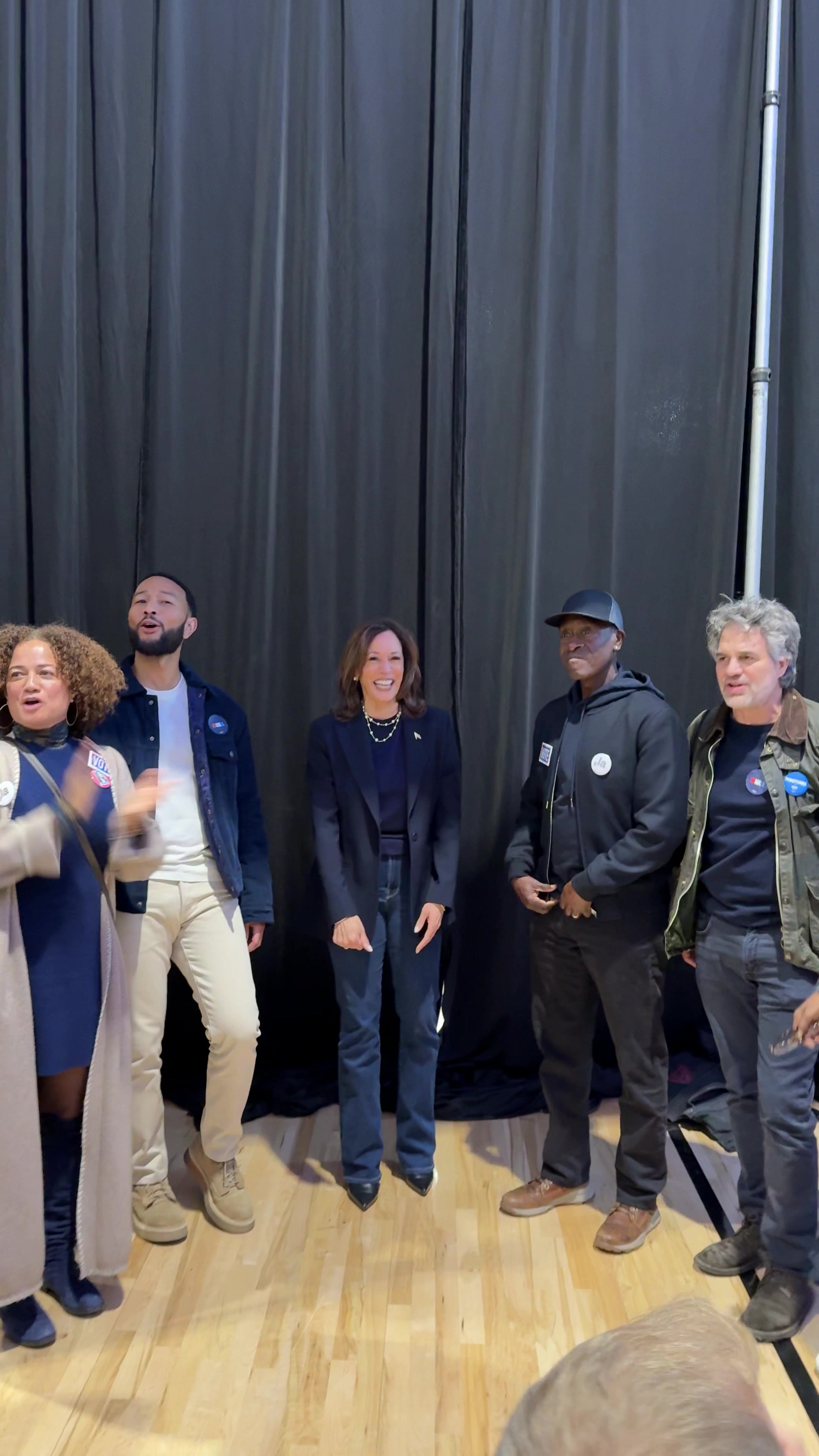 The cover image shows a group of six people standing together indoors. The lighting suggests the photo was taken backstage or in a similar setting. The background consists of dark, vertically draped curtains. The people are diverse and are dressed in a variety of styles, suggesting they are friends or acquaintances. From left to right, the first person is a woman with curly hair, dressed in a blue dress and a long cream-colored coat, gesturing with her hands. Beside her a man in a white t-shirt, blue denim jacket and light-colored pants is singing and posing. Next to him is a woman in a navy-blue suit and blue jeans. Next, there is a man wearing a black cap, black jacket and dark pants. Finally, is the man in a jacket, dark blue t-shirt and jeans. The floor is made of light-colored wooden planks, implying an event venue or backstage area. 