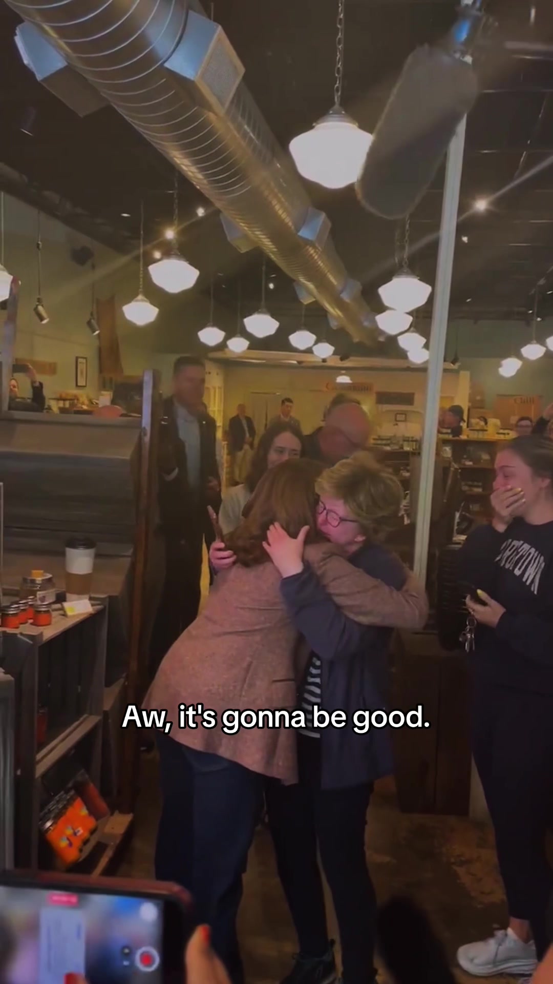 A candid indoor scene capturing a group of people embracing in an emotional moment. The setting appears to be a restaurant or business interior, with a mix of people expressing various emotions; joy, surprise, and comfort. The lighting is warm, creating an atmosphere of togetherness and support.