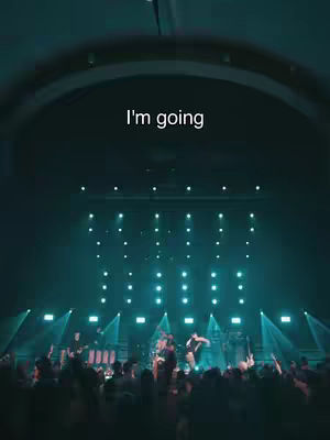A wide shot of a concert stage with a band playing. The stage is lit up with vibrant teal and blue lights. In the foreground, there is a crowd of people watching the show. A text overlay reads \"I'm going\" at the top.