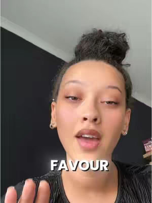 A close-up shot of a woman with dark hair pulled back in a bun. She is looking at the camera and speaking. A text overlay at the bottom of the frame reads "FAVOUR". The background behind her is dark and out of focus.