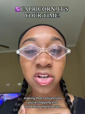 A selfie-style cover image featuring a Black woman with braided pigtails wearing diamond-shaped clear glasses. The background is a neutral-toned room. Text overlays at the top read 'CAPRICORN, IT'S YOUR TIME!'