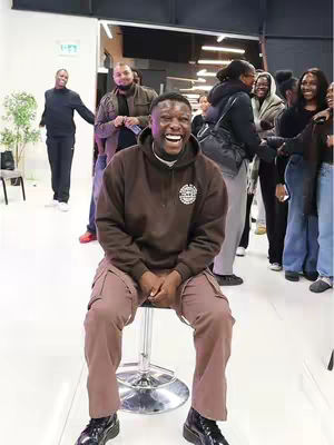 A candid image of a man laughing heartily while seated on a stool in what appears to be a youth gathering space. He is wearing a brown hoodie and matching cargo pants, with several people visible in the background. The lighting is bright, focused on the main subject.