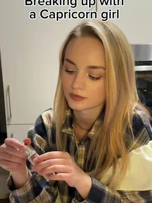 A young woman with long blonde hair, wearing a plaid shirt, is looking down while holding a lip product. She is in a kitchen setting.
