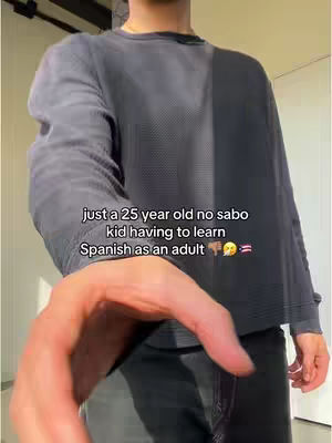 A person is shown from the waist up, wearing a dark gray long-sleeved shirt and dark pants. The person's right hand is in the foreground. The text overlay is in white, and it reads, "just a 25 year old no sabo kid having to learn Spanish as an adult"