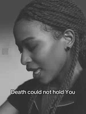 A black and white image featuring a close-up of a person's face, likely a woman, with braided hair. She is looking down with her eyes closed or nearly closed, and her mouth is slightly open as if she is singing or speaking. The text overlayed on the video reads: Death could not hold You.