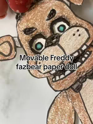 Close-up image of a handmade Freddy Fazbear paper doll with a black text overlay. The doll is brown and tan, with white eyes and light blue irises. The mouth is open in a wide grin, showing teeth. The text reads 'Movable Freddy fazbear paper doll'.