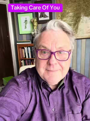 A man with gray hair and purple-framed glasses is centered in a selfie, in front of a room with a bookshelf and framed pictures. He's wearing a purple button-down shirt. Above, a text overlay reads 'Taking Care Of You' in a pink box.
