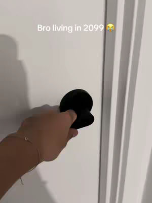 A hand holding a black doorknob on a white door. Text overlay says 'Bro living in 2099' with a smiling emoji.