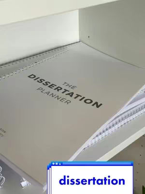 The image showcases a dissertation planner lying open in a white cabinet or shelf. The topmost part of the planner, which is facing the viewer, has the words 'THE DISSERTATION PLANNER' in large, black, clear font. Below, in a corner of the image, a blue and white box with the word 'dissertation' is overlaid.
