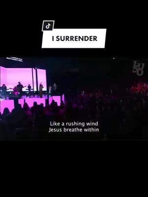 The cover image shows a live worship performance on stage. The stage is brightly lit with pink and white lights, and the audience is visible in silhouette in the foreground. Text overlay reads: “I SURRENDER”.