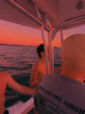 A boat at sunset with several people on board. The sky is a gradient of pinks and oranges. Several people, partially visible, stand on the boat deck. One shirtless individual is turned towards the camera. Another person is facing the other way.