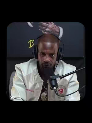 The image shows a man in a white jacket with a logo and text, sitting in what appears to be a studio setting, wearing headphones and speaking into a microphone. Behind him is a blurred background with text and a logo.