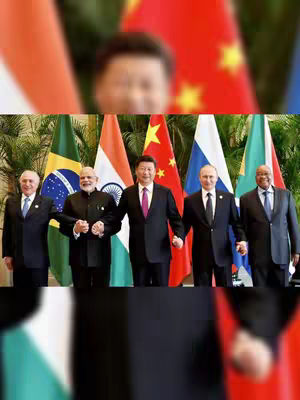A group of five world leaders stands together holding hands in front of their respective national flags. The individuals are in suits. The flags include those of Brazil, India, China, Russia, and South Africa. The background is blurred, but appears to be an indoor setting.