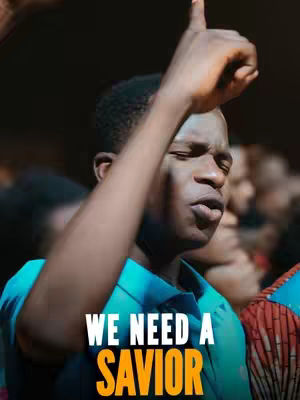 A medium shot featuring a young man, seen from the chest up, with a slightly blurred background of other people. He has his eyes closed and his face tilted upwards, as if in prayer or worship, against a fairly dark background. His right arm is raised, with the index finger pointing upward. The man is wearing a blue collared shirt, and the image has the superimposed text 'WE NEED A SAVIOR' in white with the word 'SAVIOR' in a contrasting orange color at bottom.