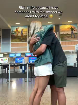 A couple is embracing tightly in an airport or similar public space. The woman, with blonde hair and wearing a white skirt and a green hoodie, wraps her arms around a person whose back is towards the camera. The background includes blurred murals and signs, set against a neutral backdrop.