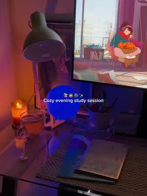 The image shows a cozy evening study setup. A desk with a book, notepad and a glass of drink are the focus. A lamp is on. In the background, a monitor displays a cartoon scene. There is also a candle.