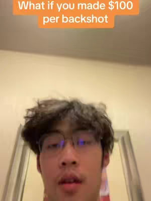 A close-up of a young man with curly hair and glasses. The image is lit with warm, natural light. Above the man's head, a text overlay in orange reads: 'What if you made $100 per backshot'. The background is a light, neutral color, suggesting a domestic or indoor setting. The man is looking towards the camera.