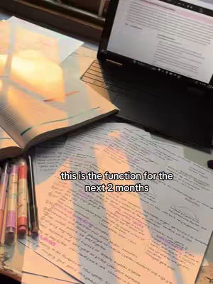 An overhead shot of a desk covered in study materials. An open textbook is toward the left, with colorful pens and pencils in front of it. Papers covered in handwritten notes are spread across the desk. A laptop sits on the right, with the screen displaying text. Soft light and shadows create a warm atmosphere.