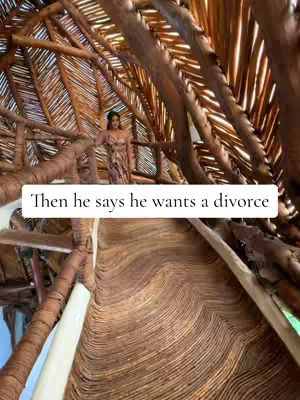 The image shows a woman in a dress standing on a walkway made of wooden planks. She is inside a structure with a thatched roof and wooden support beams. White text overlayed reads, 'Then he says he wants a divorce.'