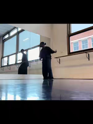 In a brightly lit room, a person dances near a large window. The floor reflects the light, and a mirror shows a blurred reflection. The dancer is wearing dark loose clothing, and there's architecture visible outside the windows.