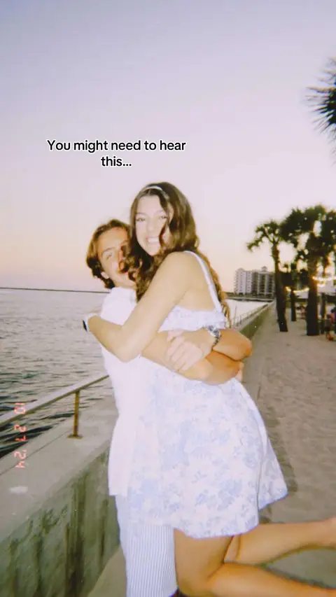 A young couple embraces on a beach at sunset, with the woman smiling and the man behind her. The background includes the ocean, a pier, and some palm trees.