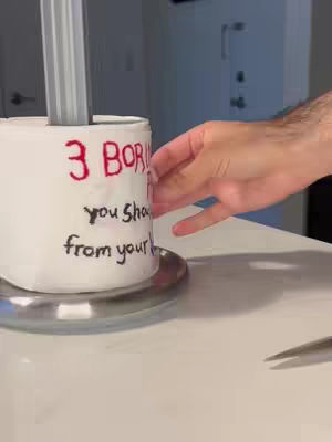 The cover image shows a white cylindrical object, possibly a paper towel roll or similar, with text written on it. The text is in a handwritten style, with some words in black and others in red. A hand is reaching towards the object next to a metallic tray sitting on a white surface where a piece of metal is leaning.