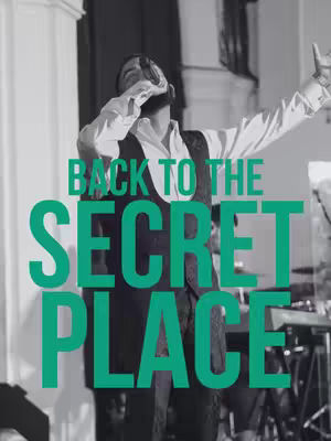A man wearing clothes and holding up a mic stands center stage, with the overlaid text 'BACK TO THE SECRET PLACE' in bright green. The background is a softly lit room with additional elements visible, all in grayscale.