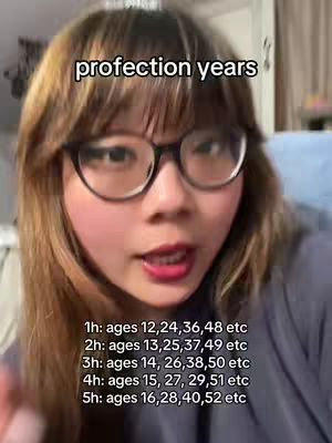 A selfie-style shot featuring a woman with glasses, set slightly off-center. She has light skin, dark hair with bangs, and is wearing glasses. The background is blurred, suggesting an indoor setting.