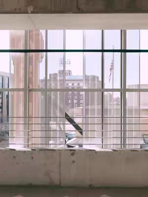 The image captures a view from a high vantage point, looking through large, divided windows and a metal railing at an urban cityscape. The foreground consists of the lower portions of windows and a concrete ledge. Beyond the window, several buildings in a muted color palette are visible, including a tall, light-colored structure and a building with a distinct sign.