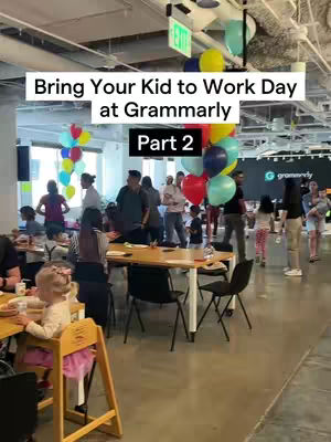 The image captures a vibrant scene during a bring-your-kid-to-work day. The composition is a horizontal shot taken in what appears to be a large office space. The scene is filled with people, children, and colorful decorations. Balloons in various colors, including red, blue, yellow, and green, are clustered together, possibly for decorative purposes. The setting includes tables and chairs, with some people seated and others standing. There is a sense of energy and activity. A young child, possibly a toddler, is seated at a table. Overall, the scene portrays a lively and positive atmosphere of a company event.