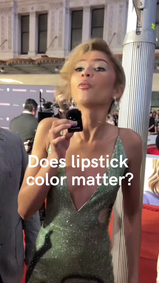 The image shows a woman speaking into a microphone. She's wearing a sparkling green dress and has blonde hair. Overlayed on the image is text that asks, "Does lipstick color matter?" The background is blurred, but suggests an event or red carpet setting, with other people visible and a tall, metallic structure.