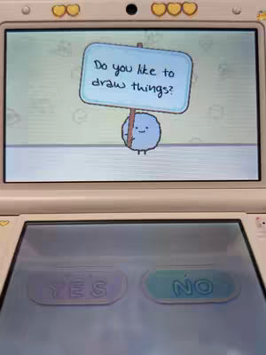 The image shows a 3DS console displaying a game screen. The top screen of the console shows a blue creature holding a sign that says, "Do you like to draw things?". Below that, the bottom screen of the console shows the options "YES" and "NO".