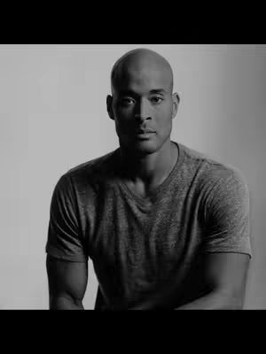 A black and white photo features a man, centered in the frame, gazing directly at the camera. He has a bald head, a serious expression, and a neutral background. He's wearing a simple grey t-shirt.