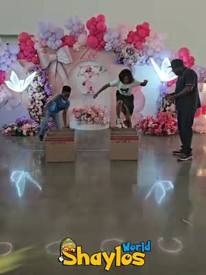A colorful birthday party scene with kids jumping over cardboard boxes in front of a decorated backdrop. Balloons and floral arrangements frame the setup.
