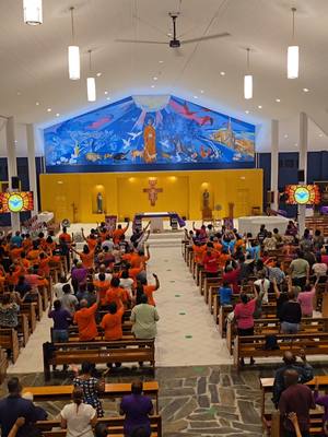 An interior shot of a church during a religious event. The image is brightly lit, featuring rows of pews filled with people, most wearing orange shirts. A large mural adorns the wall behind the altar, and the overall composition emphasizes the communal gathering. The perspective is from the back of the church towards the front.