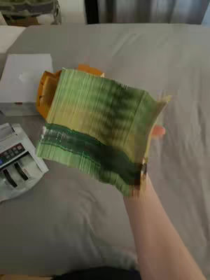 A first-person view shows a hand holding a stack of green Canadian currency. The person's arm and hand are visible, with the bills fanned out. Other objects like a white box, and a money counter can be seen on the background.