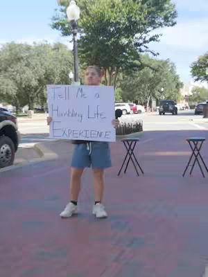A person holds a white sign with black handwritten text on a brick-paved street. They stand in front of a backdrop of trees, a street lamp, and parked cars. Two small black stools are positioned to the right.