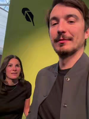 The image features a man in the foreground and a woman in the background. The man is wearing a dark jacket over a black shirt. The woman is wearing a black shirt and a necklace. The background has a green wall with a black leaf symbol.