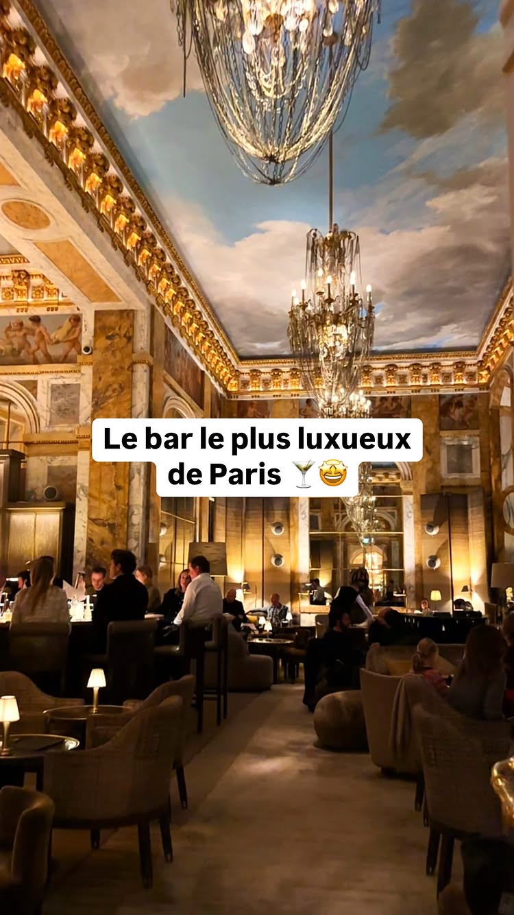 An interior shot of a luxurious bar with ornate ceiling with painted sky scenes, and large chandeliers. Tables and seating arrangements filled with patrons are visible, and a bar area.