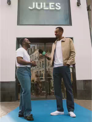 Two men standing outside a store entrance, one holding a microphone and interviewing the other. The store has a large sign that says 'JULES'.
