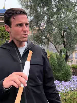 A man is holding a churro with a white paper wrap. The image is set outdoors in front of a garden with purple flowers and a topiary. The man is looking to the left. The scene is well-lit, showcasing a clear, pleasant environment.