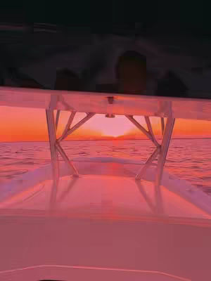 A first-person perspective from inside a boat looking out at a golden sunset reflecting on the water. The boat has a white frame in the middle of the image with a person out of focus in the distance.