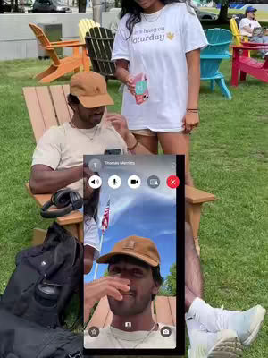 The cover image captures a video call scene within a casual outdoor setting. A man, wearing a brown cap and a beige t-shirt, is seated on a wooden chair, with headphones around his neck. He is holding a phone and is interacting with someone on a video call. A woman in a white t-shirt, with text on it, and shorts is standing next to him. The background shows green grass, wooden leisure chairs, and part of a building. Additional elements include a black bag and an American flag on the phone screen.