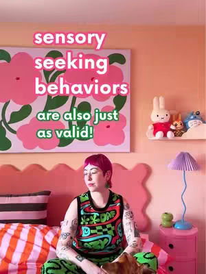 A person with pink hair and tattoos sits on a bed with a colorful background featuring a large floral painting and decorative items. The hook text reads, 'sensory seeking behaviors are also just as valid!'