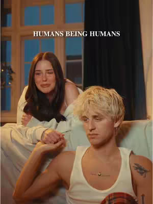 A cover image of a video featuring a man and a woman in a room. The man is in the foreground with a neutral expression, wearing a white tank top and has tattoos on his arm and chest. The woman is in the background lying on the bed with a sad expression, dressed in a white sweater. The text 'HUMANS BEING HUMANS' is present at the top.