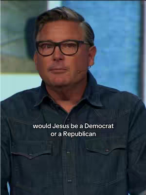 A middle-aged man with short, graying hair and glasses stands in front of a blue-toned background. He's wearing a denim shirt. Text overlaid near the bottom of the image reads, 'Would Jesus be a Democrat or a Republican'.